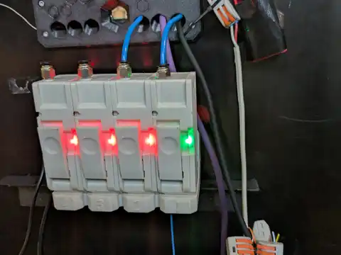 A grey four-channel module mounted on a black panel, its channels numbered 4, 3, 2 and 1, three showing red indicator lights and one green. Blue and purple tubing runs into metal push-fit fittings along the top edge. Above it a black 3D-printed plate with unused hexagonal and round recesses and a single gold mirrored hexagon. To the right, orange lever-nut connectors and a black-taped assembly with a red circuit board. Black honeycomb mesh with blue panelling behind fills the top of the frame.