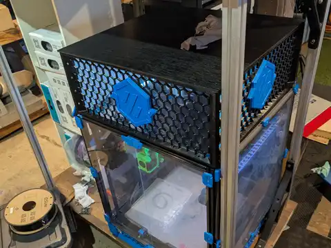 The Voron 3D-printer enclosure on a rolling cart, its black frame carrying honeycomb-mesh side panels and hex Voron badges, clear front doors closed over the print bed; filament spool boxes and shelving fill the workshop background.