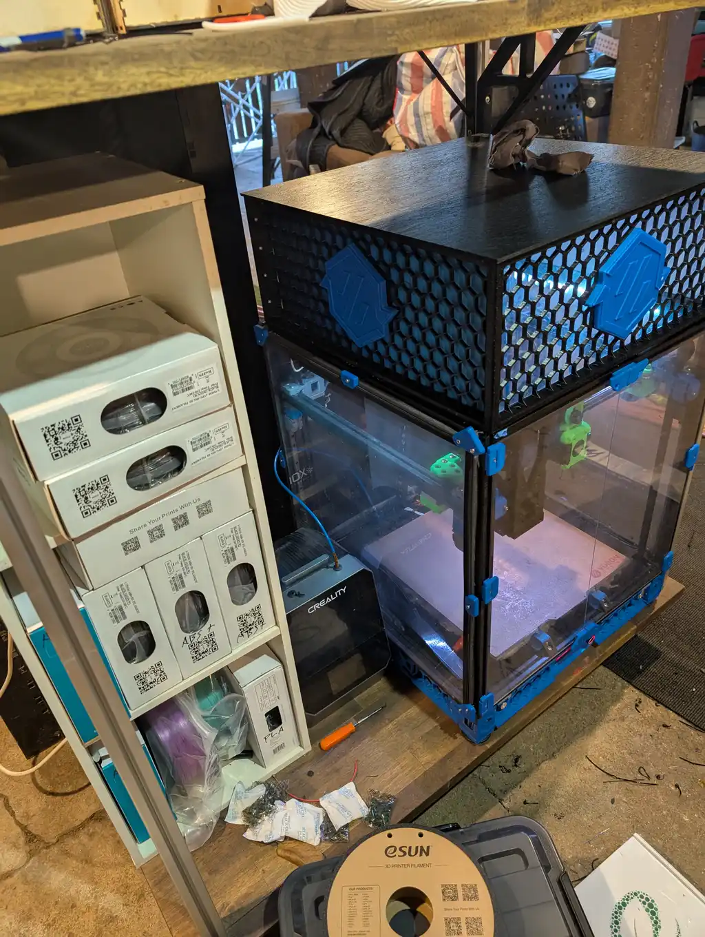 The same enclosure viewed from its other side, showing the mesh top panel, blue printed door latches and the clear front doors, with a Creality-branded printer and stacked filament boxes on the shelf beside it.