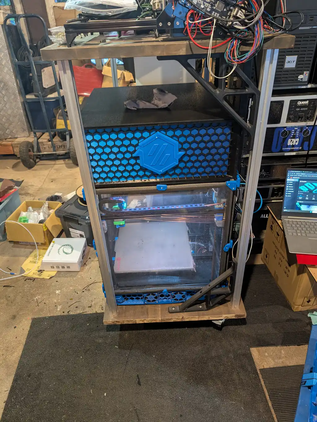 The enclosure's front corner with its clear door open and the print bed and gantry visible inside; rack-mounted audio amplifiers sit on the shelf to the right, and at the edge of frame a laptop screen shows a printer control dashboard.