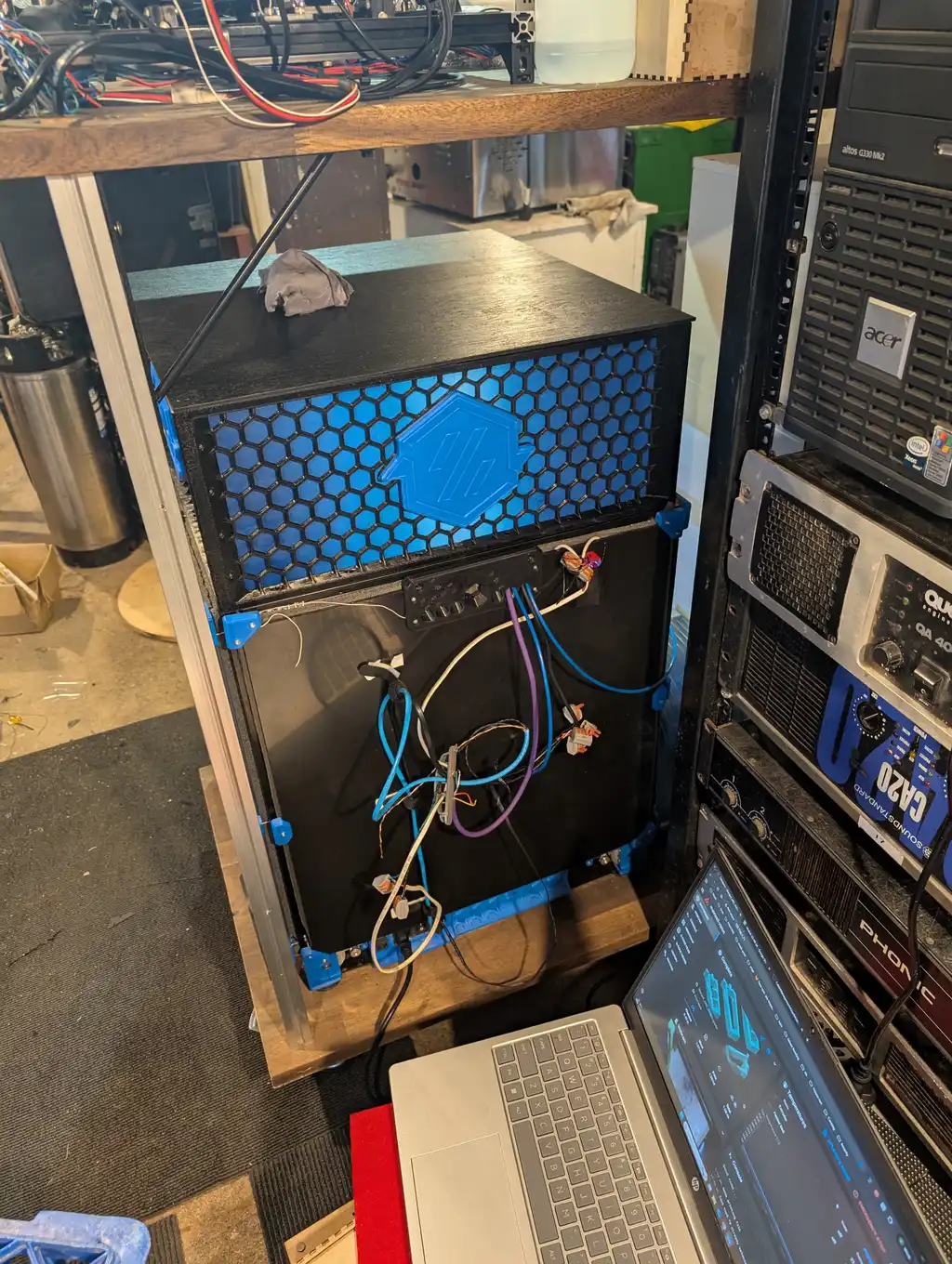 The enclosure's open back panel, exposing a wiring loom of blue, purple and white cables running to its control boards; in the foreground a laptop's screen shows a printer control dashboard, with rack-mounted audio equipment visible behind it.