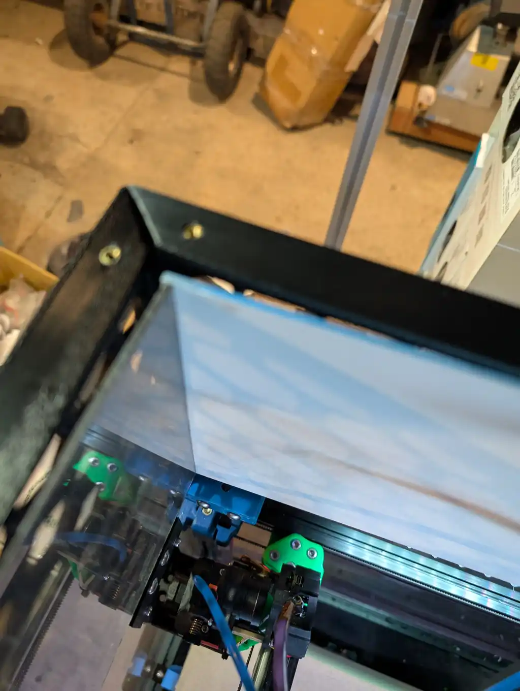 A close-up beneath the enclosure's gantry, showing a curved translucent diffuser panel, the black frame corner, a drive belt and green 3D-printed mounting parts, with a wheeled cart blurred in the background.