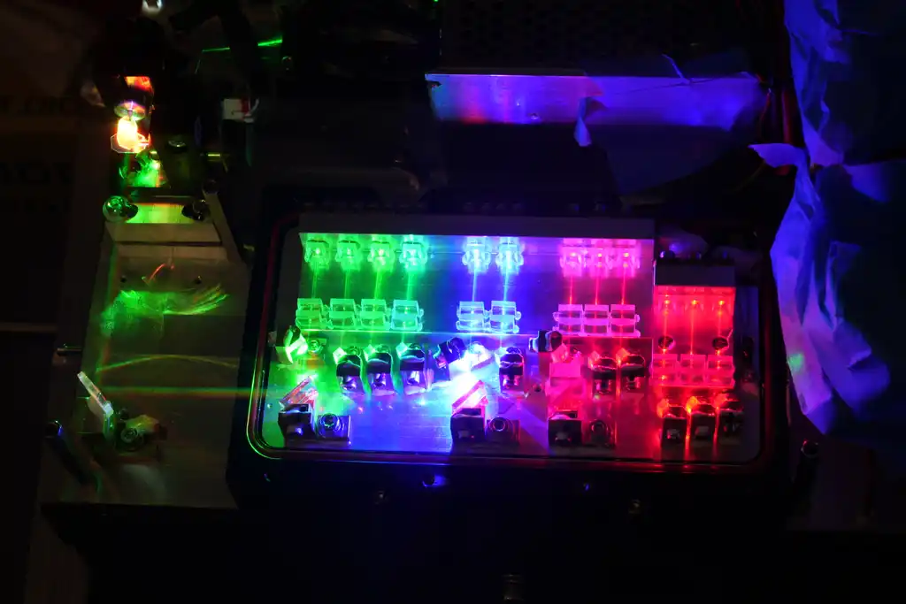 A closer, low-light view of the laser-diode projector rig's array lit and running, showing the individual diode modules in green, blue-white, magenta and red mounted along the heatsink bar, with further optics and a coiled cable to the left.