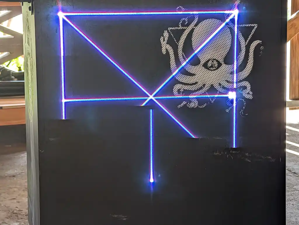 A finished laser projection from the laser-diode projector rig on an outdoor wall at night: a rectangle and crossing diagonal lines in blue and magenta light, with a stencil-cut octopus graphic caught in the beam.